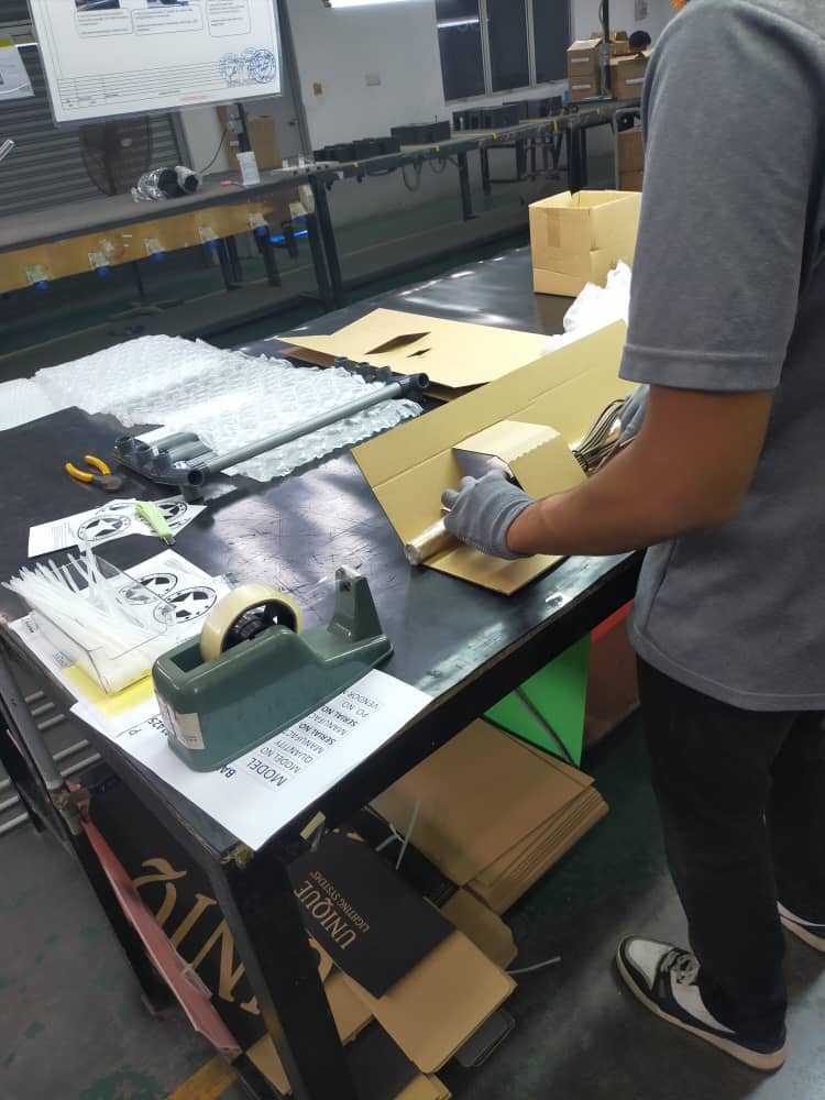 A factory worker carefully packaging a brass landscape lighting fixture for shipment, demonstrating Amat Sinar Lighting's commitment to quality and product protection.
