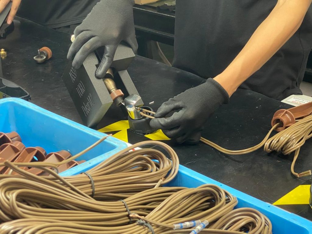 Technician in black gloves assembling brass landscape lighting components on a black workbench with spools of wire in a blue bin.