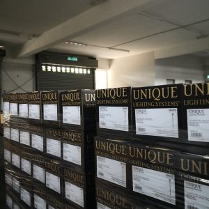 Pallets stacked with boxes of UNIQUE brand lighting systems, primarily featuring brass path lights, wrapped in plastic for shipping.