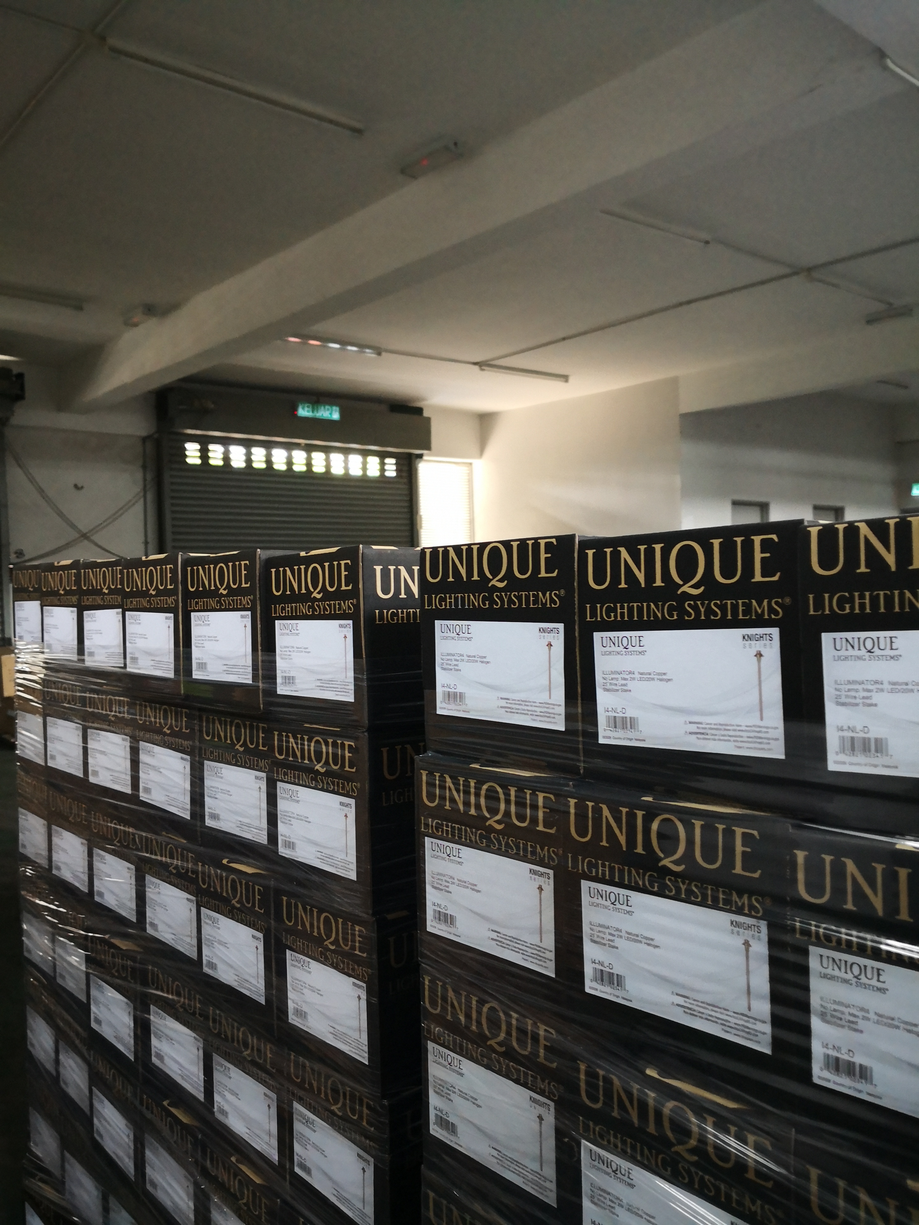 Pallets stacked with boxes of UNIQUE brand lighting systems, primarily featuring brass path lights, wrapped in plastic for shipping.