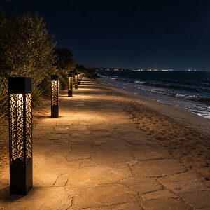 Black geometric bollard lights illuminating a stone pathway along a sandy beach at night, with the ocean and moon visible in the background.