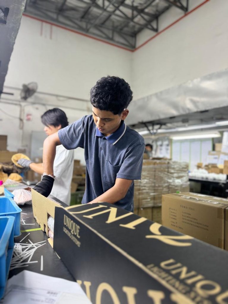 Young man in a blue polo shirt and black gloves working on a conveyor belt, assembling brass landscape lighting fixtures inside a factory.