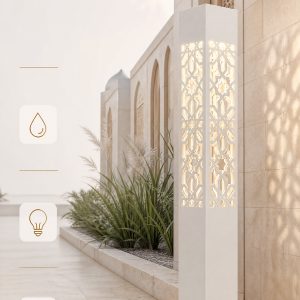 A modern white geometric bollard light with a laser-cut Arabic pattern, illuminating a pathway next to a light stone building and lush green plants.
