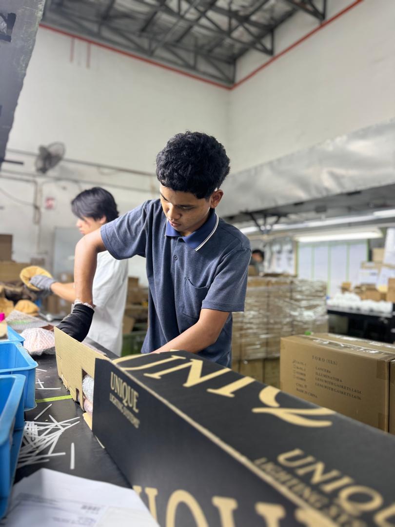 Young man in a blue polo shirt and black gloves working on a conveyor belt, assembling brass landscape lighting fixtures inside a factory.